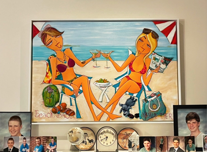 Beach Babes hanging in a white frame above a cabinet of family photographs and clocks in Eric's home