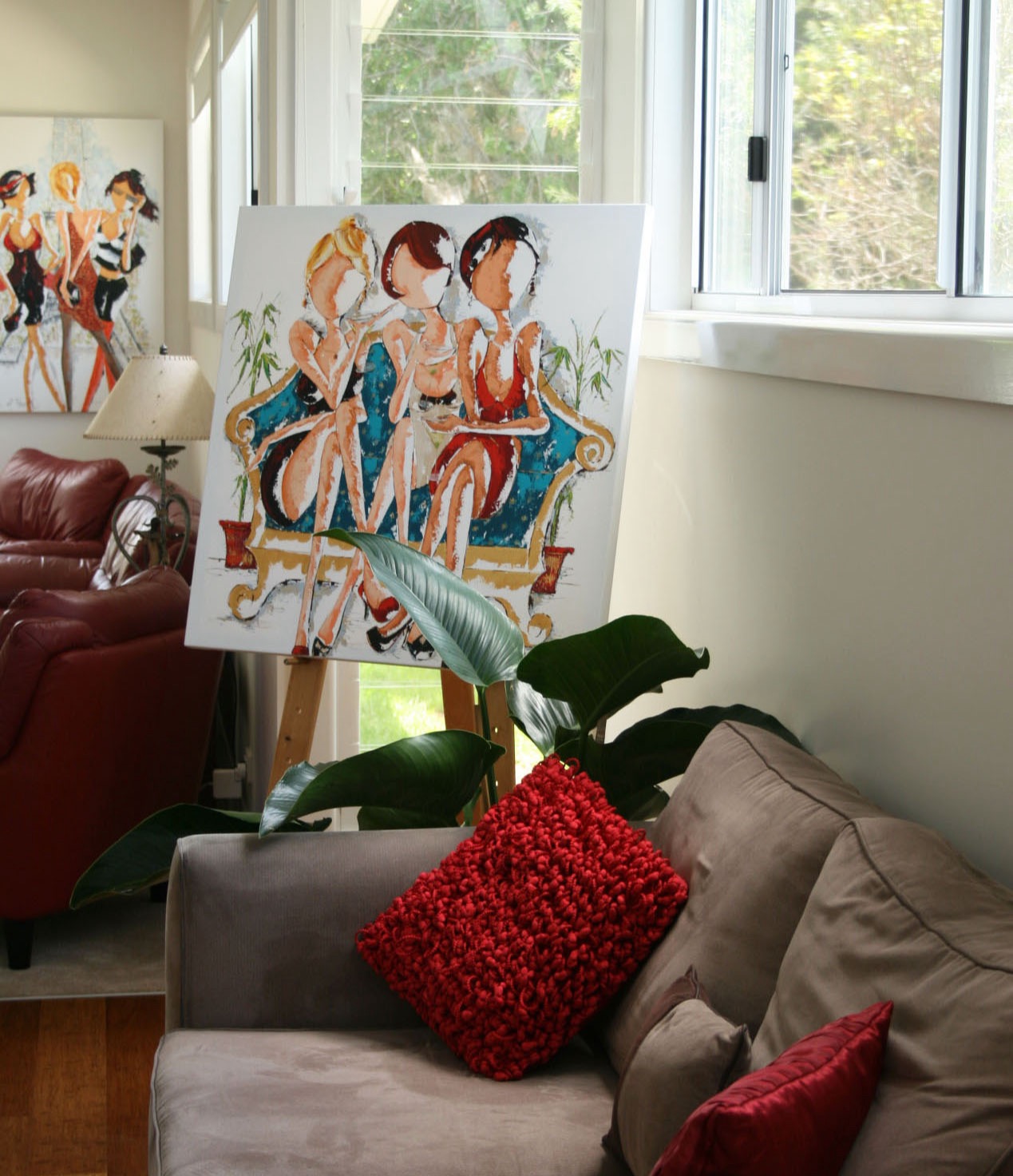 The original Oh My Goodness on an easel in a sunroom, beside a grey sofa with a red textured cushion and a large philodendron